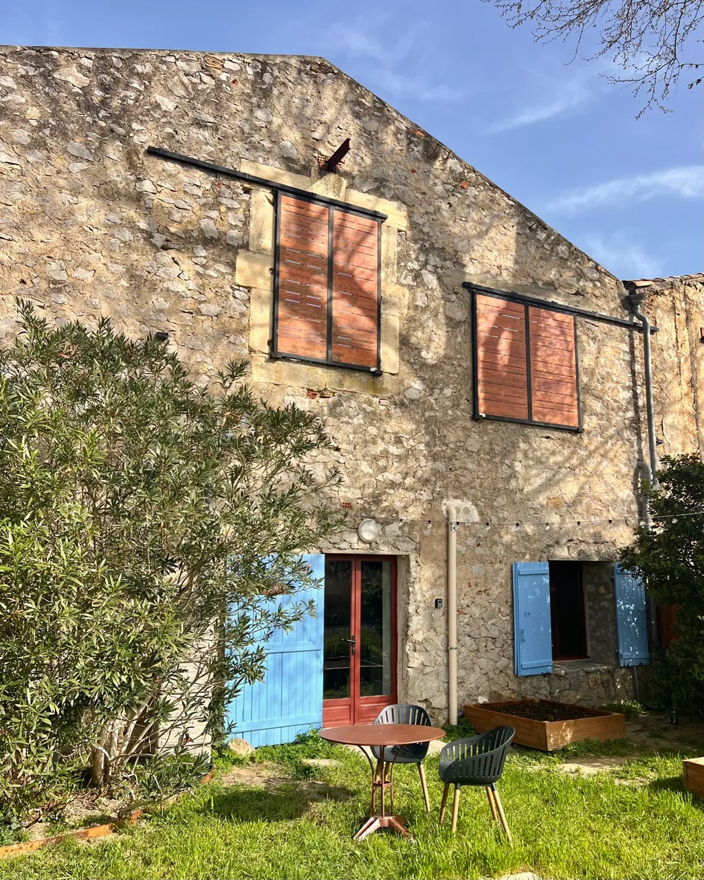 Die Natursteinfassade des Hauses mit Giebel, hellblauen Fensterläden und einem Oleander, davor ein kleiner Bistrotisch im Gras