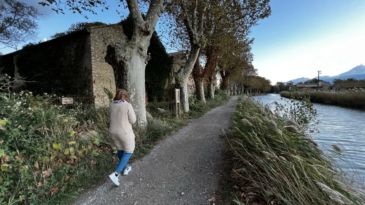 Eine Spaziergängerin auf dem Kiesweg am Kanal, links ein altes Steingebäude und eine Reihe Platanen, rechts das Wasser der Robine im Abendlicht