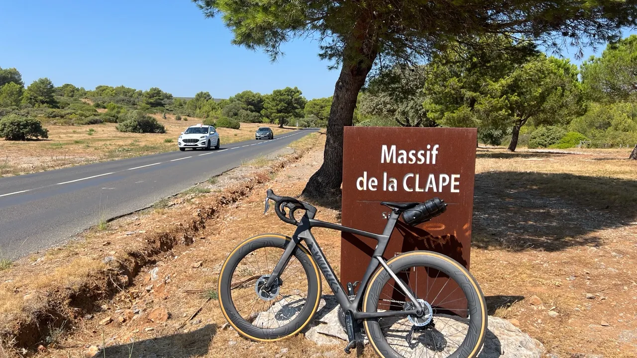 Ein schwarzes Rennrad lehnt am Cortenstahl-Schild des Massif de la Clape an der Straße durch die Garrigue
