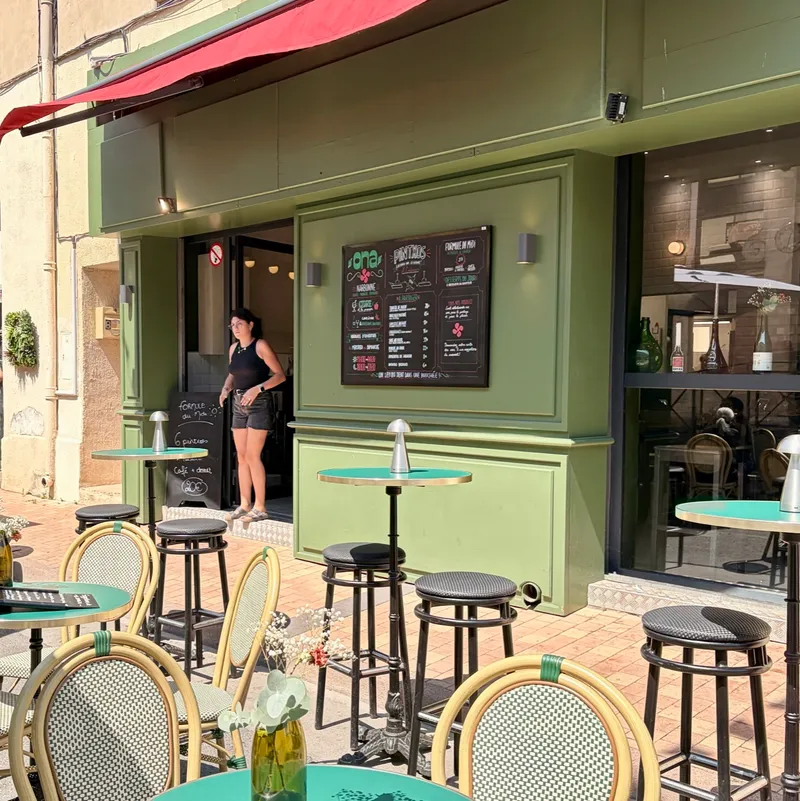 Die grüne Fassade des Ona in Narbonne mit roter Markise, Tafelmenü und türkisen Bistrotischen auf der Terrasse