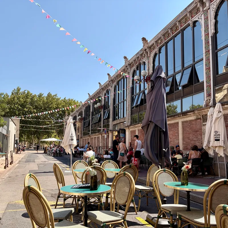 Die Terrasse des Ona mit türkisen Bistrotischen, gegenüber die verzierte Belle-Époque-Fassade der Halles de Narbonne