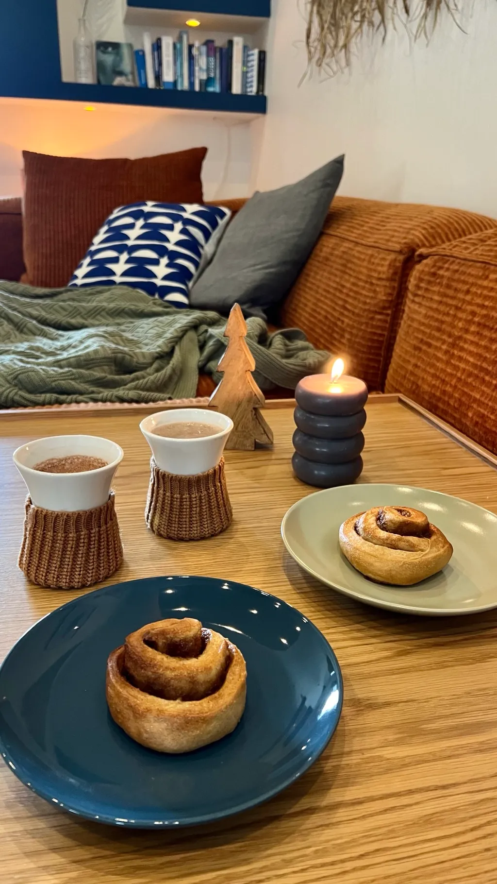 Zwei Tassen Kakao mit Strickmanschetten und Zimtschnecken auf dem Couchtisch, dahinter die Cord-Sofalandschaft mit Kissen und Wolldecke im Kerzenlicht