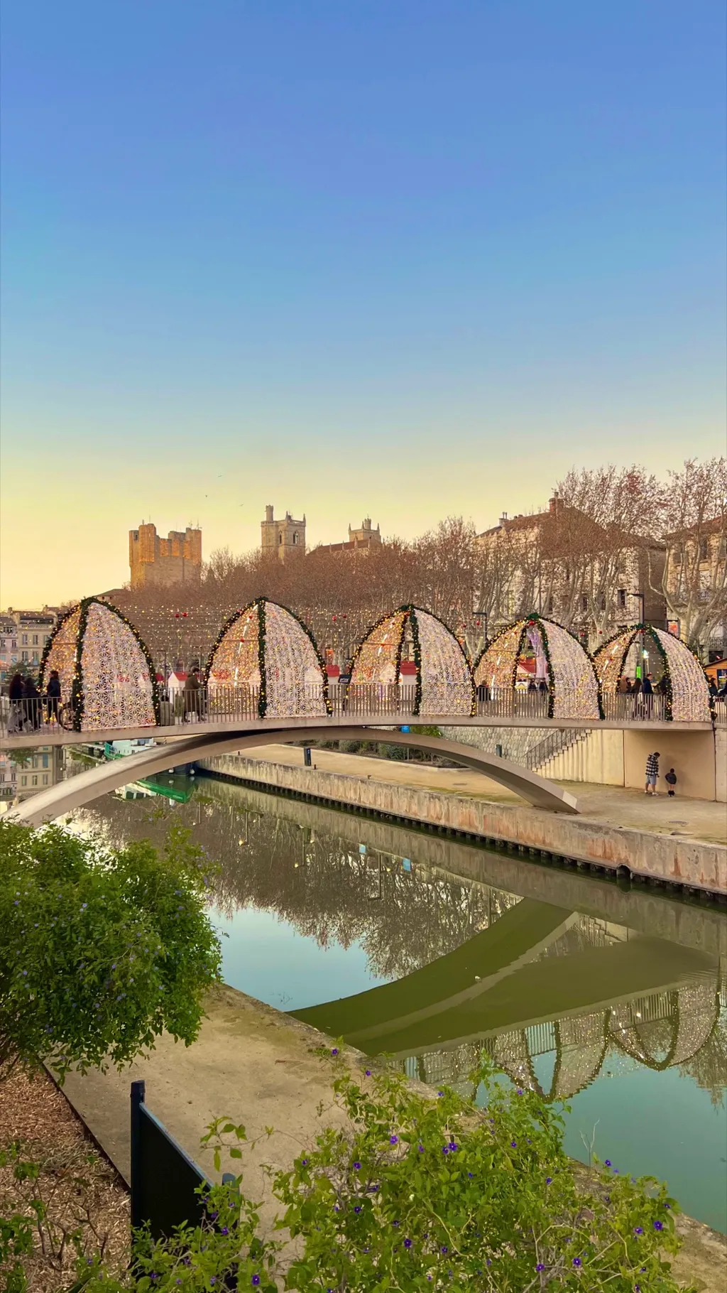 Leuchtende Lichtbögen auf der Passerelle über den Canal de la Robine, dahinter Donjon und Kathedrale in der Abenddämmerung