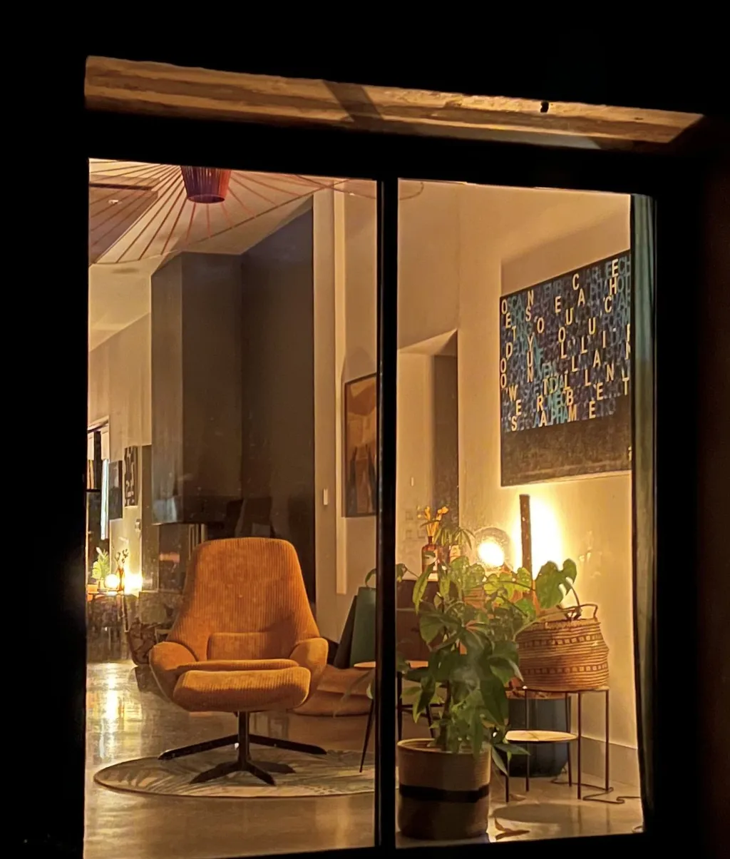 Blick von draußen durch die Terrassentür in den beleuchteten Wohnraum mit orangem Sessel, Lampe und Pflanzen