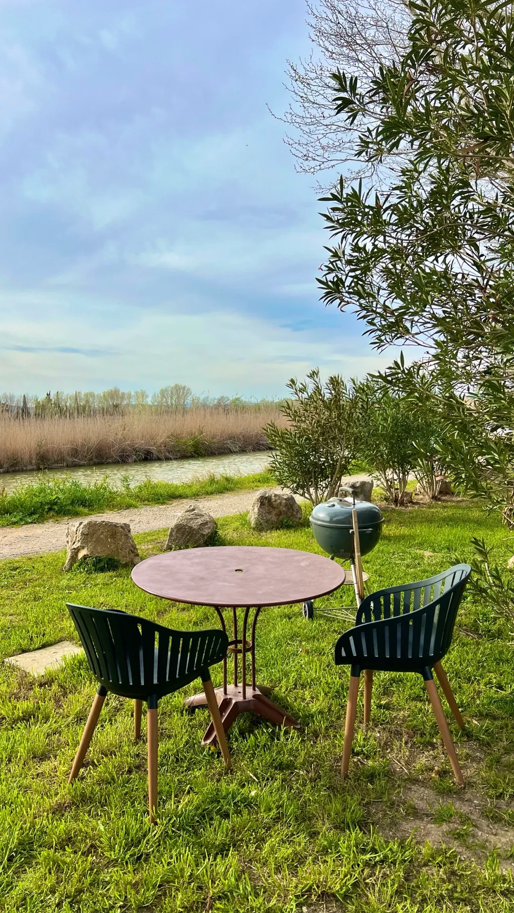 Runder Gartentisch mit Stühlen und Kugelgrill auf der Wiese am Kanal, dahinter das Schilf im Frühlingslicht