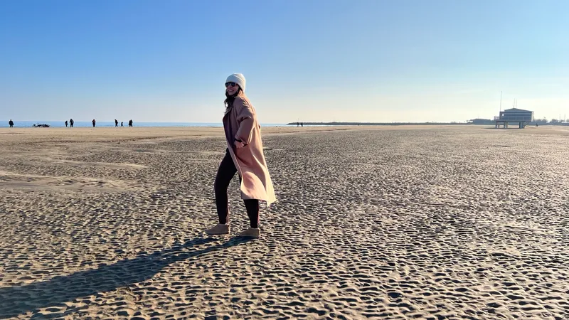 Spaziergängerin in Mantel und Mütze am fast leeren Winterstrand unter blauem Himmel