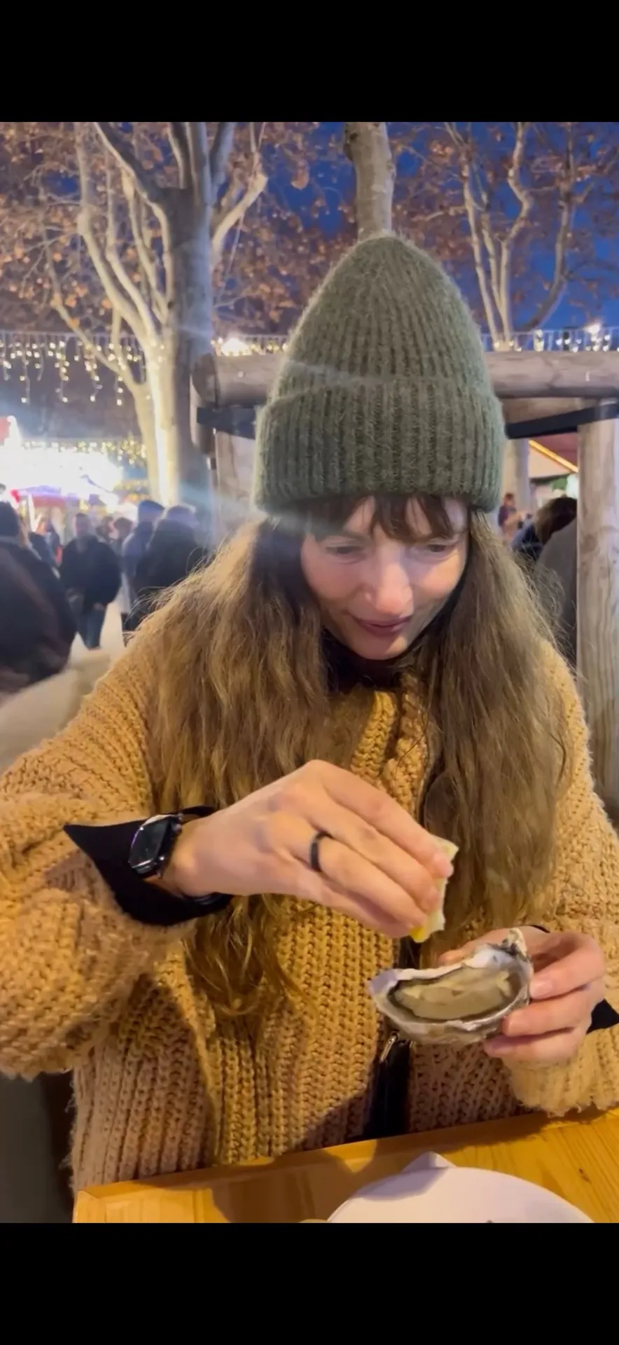 Eine Frau träufelt am Abend Zitrone über eine frische Auster, im Hintergrund die Lichter des Weihnachtsmarkts