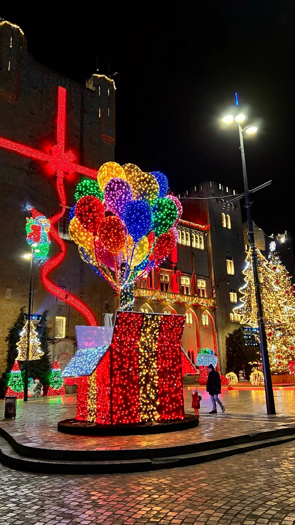 Die riesige leuchtende Geschenkschleife am Rathausturm von Narbonne, davor beleuchtete Weihnachtsbäume und Lichtfiguren
