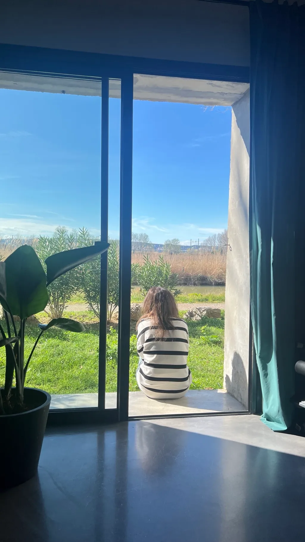 Blick durch die offene Terrassentür: eine Frau sitzt in der Märzsonne auf der Schwelle, davor Garten und Schilf am Kanal